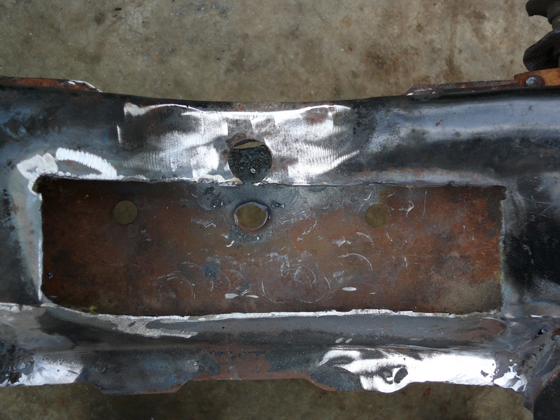 Close-up of a rusty, welded car frame section with visible circular indentations and a rectangular cutout