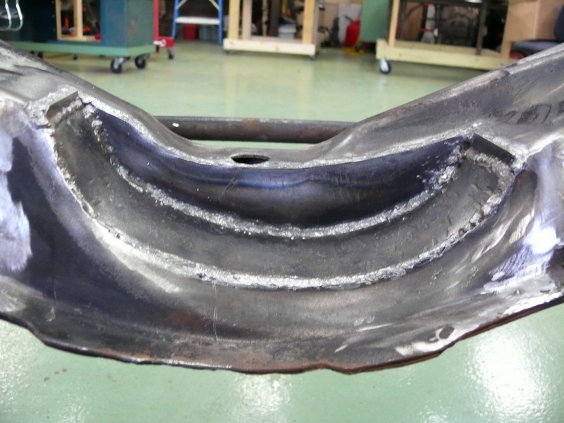 Close-up of a dark metal car frame component with visible welds and a central hole