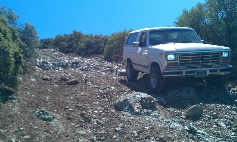 1986 Ford Bronco - A white 1986 Ford Bronco navigates a , rocky terrain under a clear blue sky.