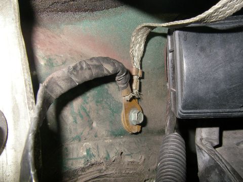 1990 Ford Bronco - braided negative ground strap and battery cable are bolted to a corroded firewall with green primer, beside a black battery case.