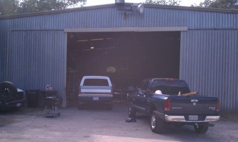 1979 Ford Bronco - A dark pickup truck is parked in front of a large metal garage with a white pickup truck inside.