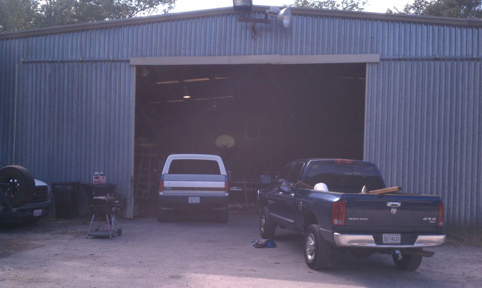 Two trucks parked outside a metal garage. One is a dark pickup, the other is a white pickup inside