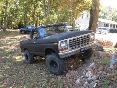1978 Ford Bronco - A black 1978 Ford Bronco is shown with a lifted suspension and large tires, parked on uneven terrain with other vehicles in the background.