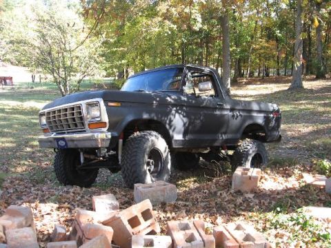 1978 Ford Bronco - This lifted 1978 Ford Bronco features large off-road tires and a dark exterior, parked amidst fallen leaves and trees.