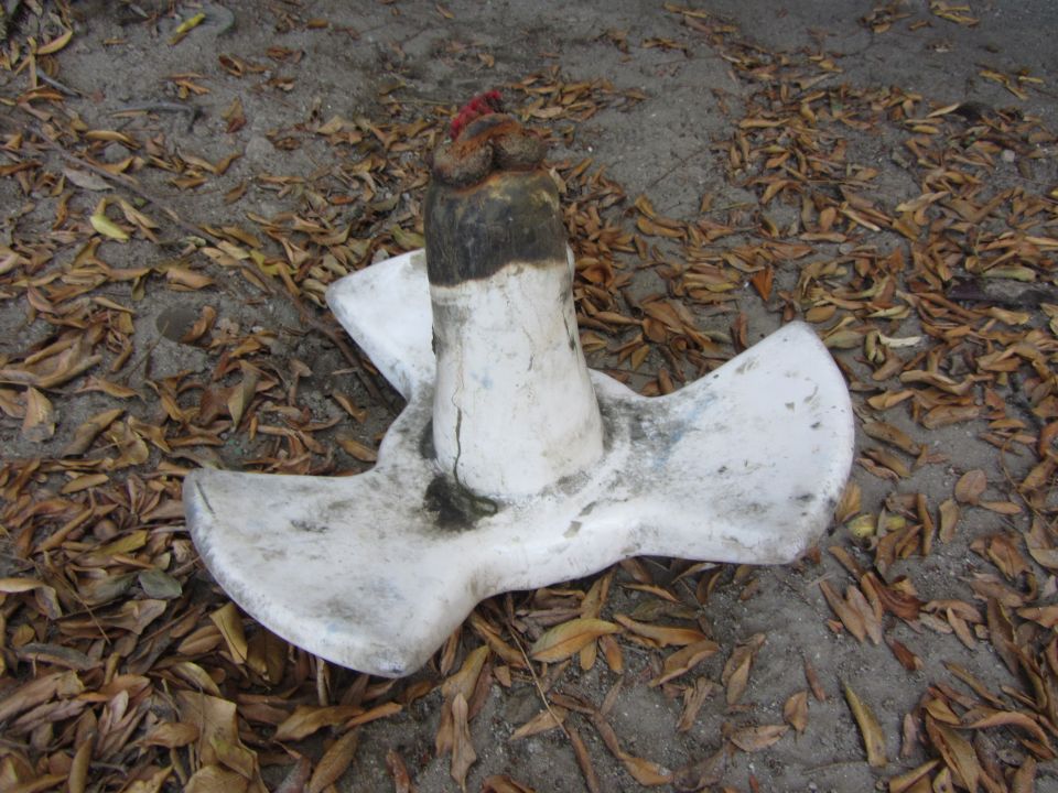 White boat anchor with a wooden post and rope on the top, lying on the ground with fallen leaves