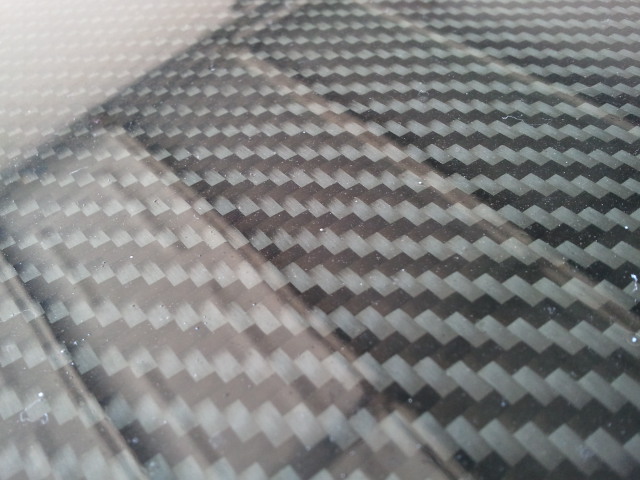 Carbon fiber pattern on a car part, with a Supermotors.net logo in the lower right corner