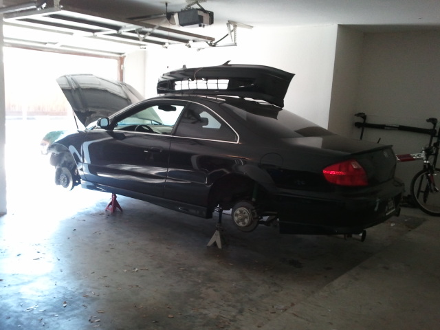 Black coupe on jack stands, hood and trunk open, with a roof box