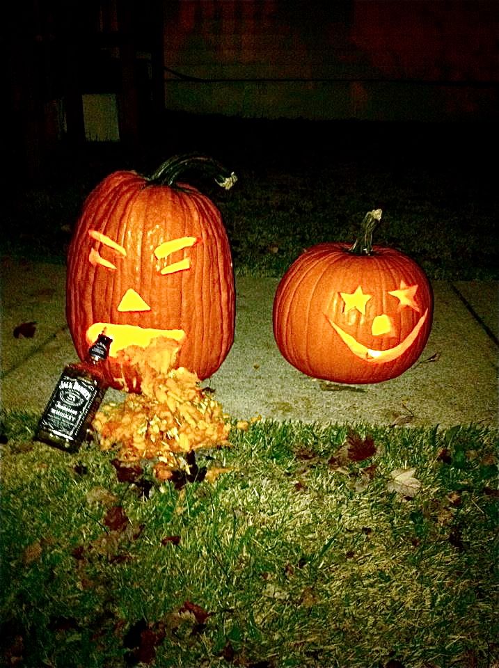 Two carved pumpkins, one with a face and spilling seeds, next to a bottle of Jack Daniels. A second pumpkin has a star-eyed face
