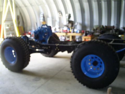 1992 Ford Bronco - The chassis of a vehicle, equipped with a blue engine and large tires with blue rims, is shown in a workshop setting.