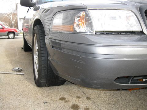 2004 Ford Crown Victoria - A gray Ford Crown Victoria with a flat tire is parked on concrete, with a red SUV in the background.