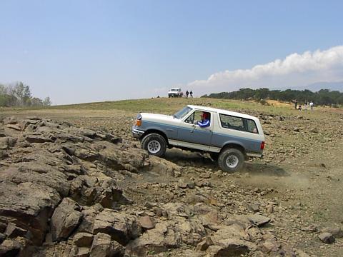 1988 Ford Bronco - A Ford Bronco navigates a challenging rocky trail under a clear blue sky with scattered clouds.