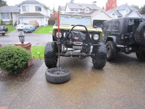 1996 Ford Bronco - Two modified Jeeps are parked in a driveway on a wet day. One has a spare tire on the ground.