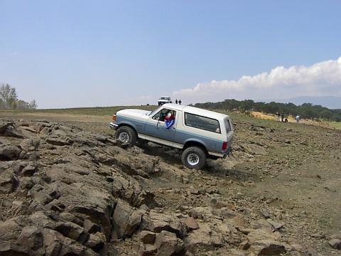 1988 Ford Bronco - A two-tone Ford Bronco navigates a challenging, rocky off-road course under a partly cloudy sky.