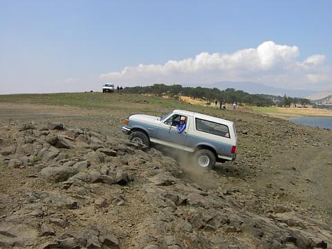 1988 Ford Bronco - A blue and white 1988 Ford Bronco navigates a rocky off-road trail with a body of water in the background.