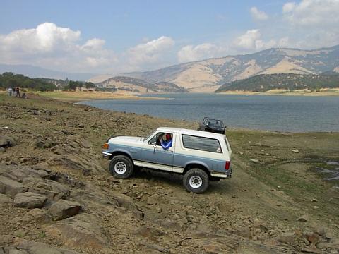 1988 Ford Bronco - Two SUVs navigate a rocky terrain beside a large body of water under a cloudy sky.