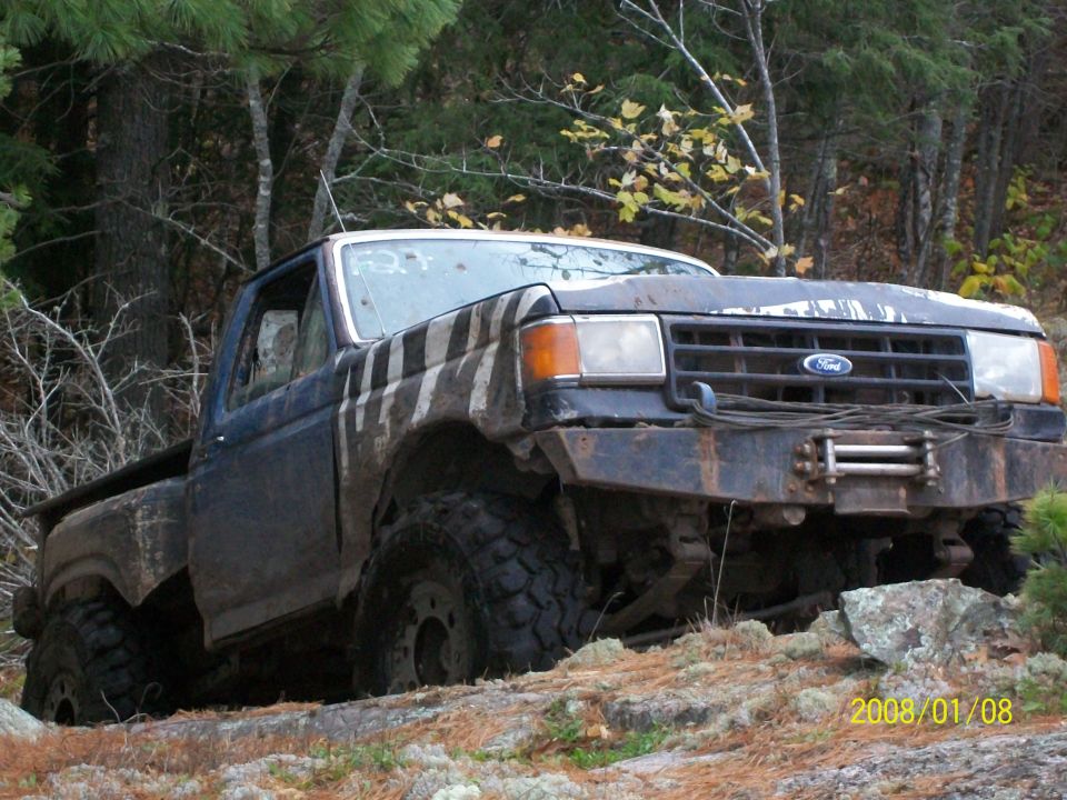 Ford pickup truck with large tires and winch on rocky terrain. The truck has a dark blue body with white racing stripes on the hood and fender