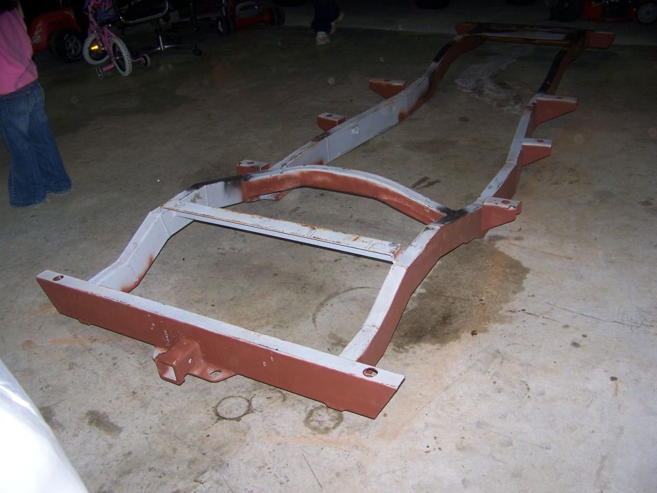 Vehicle frame with red and gray primer, resting on concrete floor