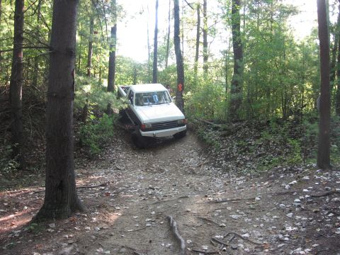 1987 Jeep Comanche - A white pickup truck navigates a wooded trail with sunlight filtering through the trees.