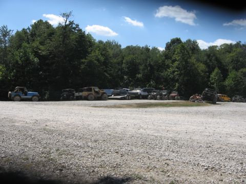 1987 Jeep Comanche - A collection of off-road vehicles are parked in a lot near a wooded area on a sunny day.