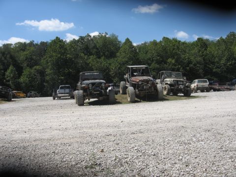 1987 Jeep Comanche - Multiple off-road vehicles, including several Jeep Wranglers, are parked on a gravel lot under a partly cloudy sky.