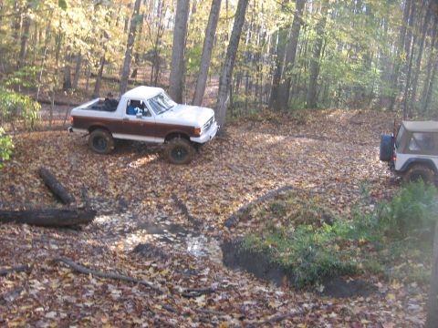 1987 Jeep Comanche - Two off-road vehicles navigate a leaf-covered trail with muddy patches in a wooded area.