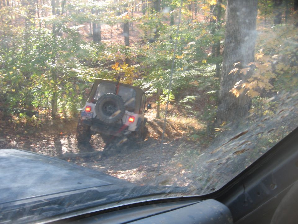 Jeep Wrangler with spare tire on rear, driving on a dirt path in a forest
