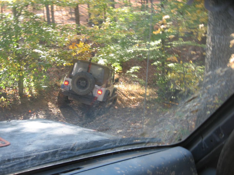 White Jeep Wrangler on wooded trail, rear view. Another vehicle in foreground