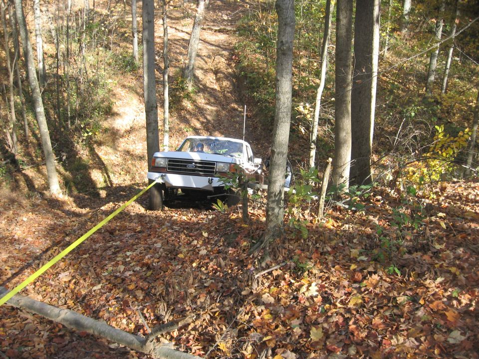 White pickup truck with a yellow tow strap, angled front view, in a wooded area with fallen leaves