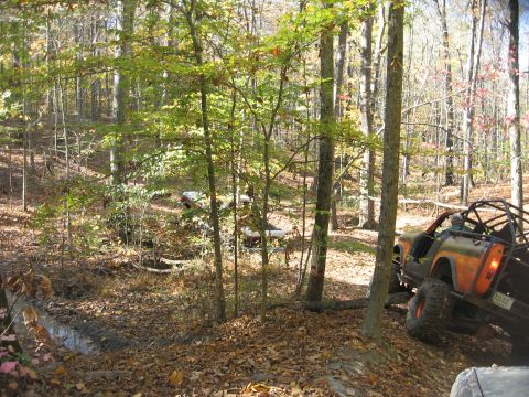 1987 Jeep Comanche - An off-road vehicle navigates a wooded trail during autumn, with another vehicle partially visible in the distance.