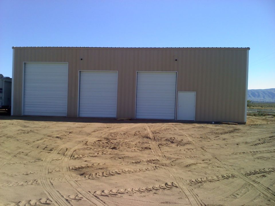 tan building with three large garage doors and a small side door, in a dirt lot with tire tracks