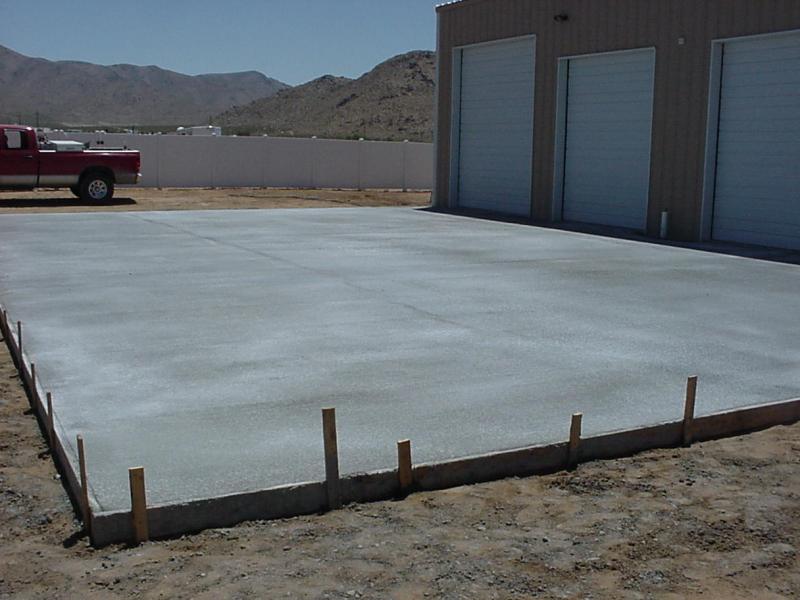 Red pickup truck parked to the left of a freshly poured concrete slab in front of a building with three garage doors