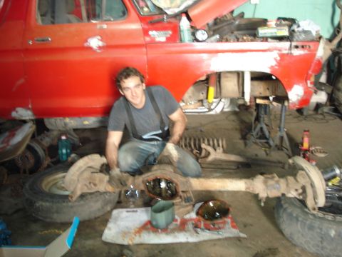 1980 Ford Bronco - A mechanic works on the front axle of a red vehicle that is raised on jack stands.