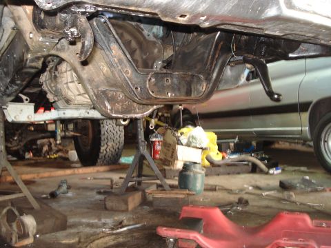 1980 Ford Bronco - The underside of a vehicle is shown on jack stands, with a second vehicle partially visible in the background, garage setting.