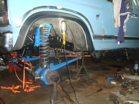 1980 Ford Bronco - Light blue SUV undergoing suspension work, showing the front axle and springs removed, with a Bronco emblem on the fender.