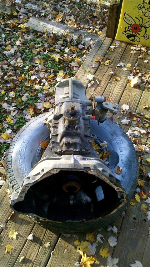 1995 Ford F-150 - A vehicle transmission rests on a tire on a wooden deck covered in autumn leaves.