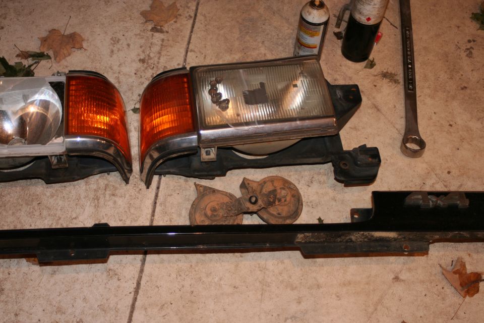 Car parts including headlights, turn signals, horns, and trim on concrete