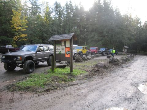 2008 Jeep Wrangler - A line of Jeeps are parked in a muddy lot near a forest, with a sign detailing park information.