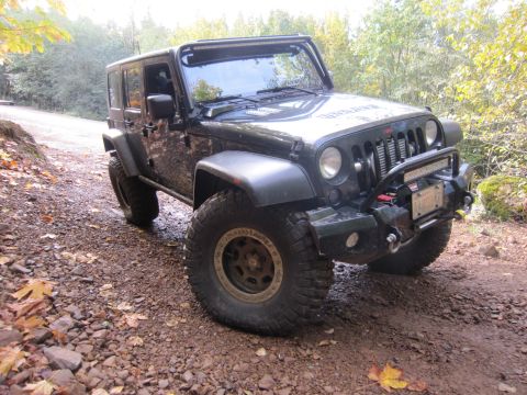 2008 Jeep Wrangler - A dark 2008 Jeep Wrangler equipped with a winch and light bar is parked on a gravelly dirt road surrounded by trees.
