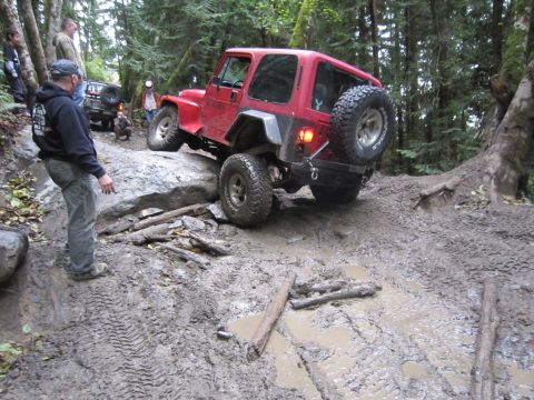 2008 Jeep Wrangler - A red Jeep Wrangler TJ navigates a challenging, muddy off-road trail surrounded by trees.