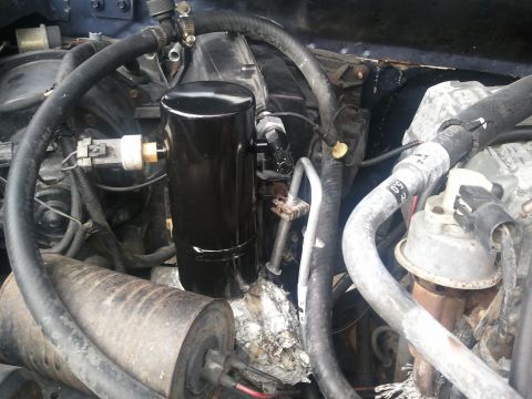 1994 Ford Bronco - under-hood view showing a black cylindrical canister, tangled vacuum hoses and wiring harnesses near a dark blue firewall.