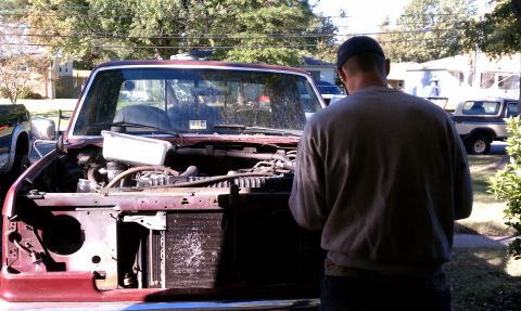 1979 Ford Bronco - A person stands in front of a red pickup truck with its hood open, revealing the engine compartment. Another vehicle is parked in the backg…