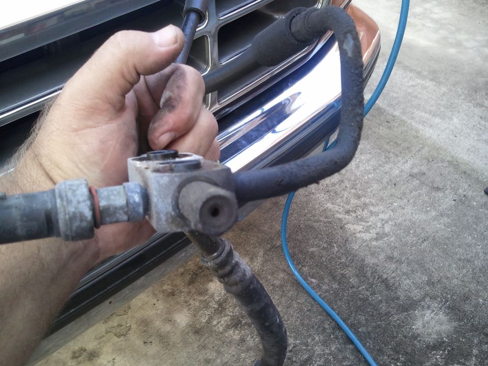 Hand holding AC hose assembly with metal fittings and black rubber hoses. Chrome grille visible in background