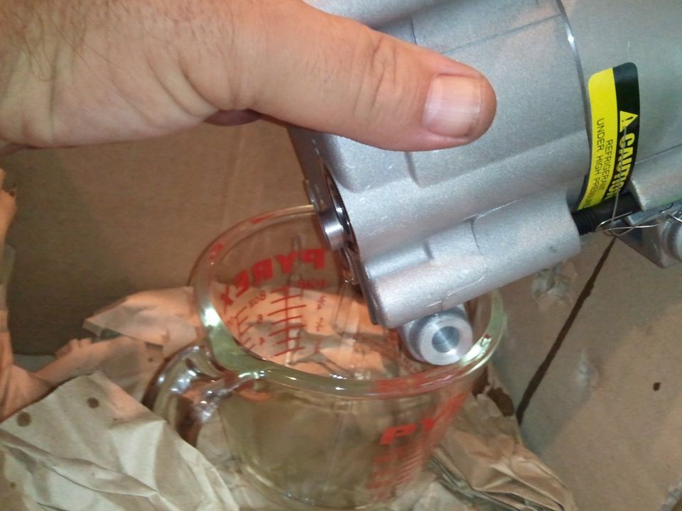 Hand holding AC compressor over Pyrex measuring cup with liquid. Yellow caution sticker visible on compressor