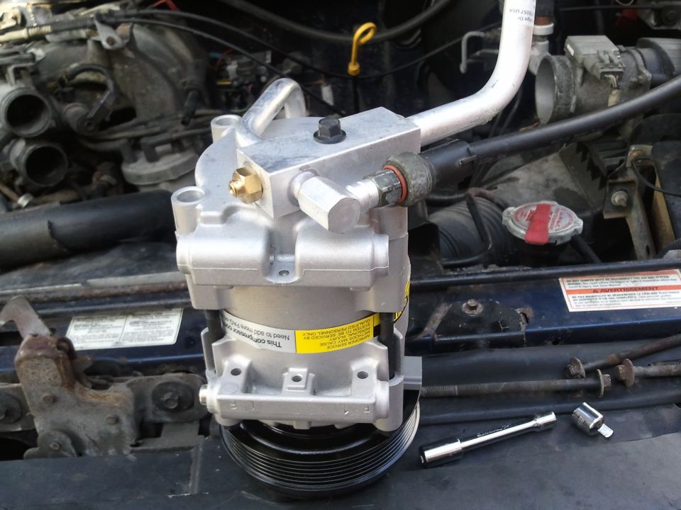 New AC compressor with yellow warning sticker, sitting in front of a vehicle's engine and radiator