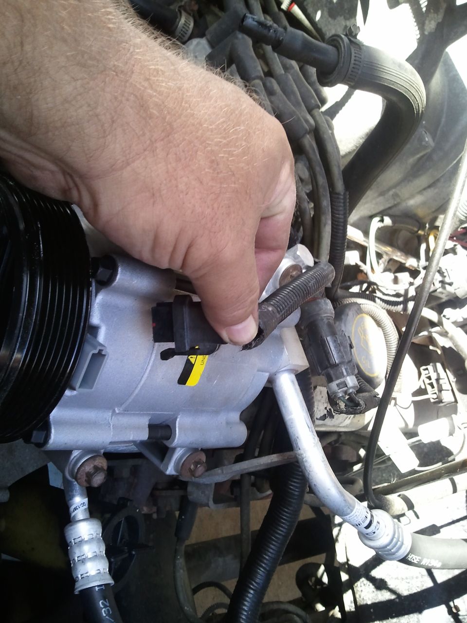 Hand connecting a wire to an AC compressor with hoses and engine parts in background