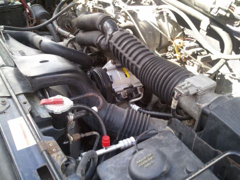 1994 Ford Bronco - Close-up view of the AC compressor and associated hoses within an engine bay. The radiator coolant reservoir is also visible.