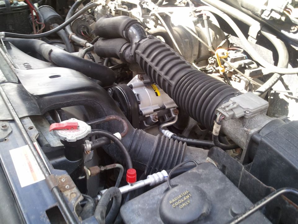 Engine bay AC compressor and hoses. Radiator coolant reservoir visible