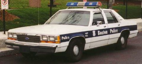 1991 Ford LTD Crown Victoria - White and blue Boston Police Ford LTD Crown Victoria patrol sedan with roof lightbar, parked beside a chain-link fence.