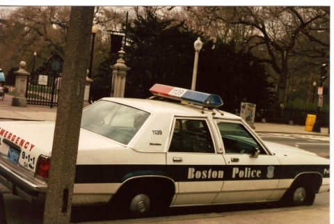 1991 Ford LTD Crown Victoria - white-and-blue Boston Police Ford LTD Crown Victoria cruiser, unit 1139, with roof lightbar parked beside an ornate park gate.