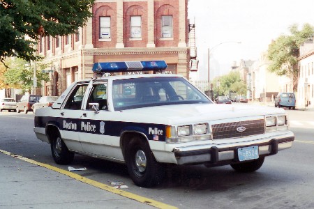 1991 Ford LTD Crown Victoria - White Ford Crown Victoria marked Boston Police cruiser with roof lightbar, parked curbside in front of a brick building.